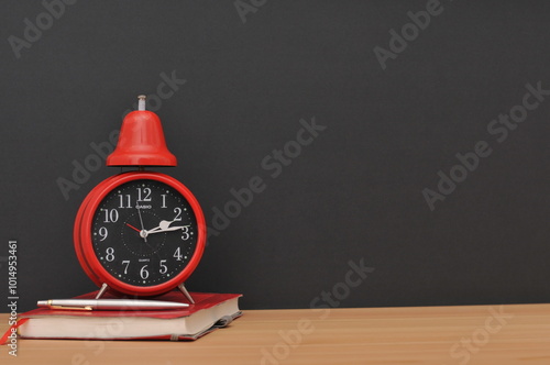 Red alarm clock on wooden table on diary with pen with black background.