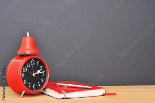 Red alarm clock on wooden table next to diary with pen. left.with black background.