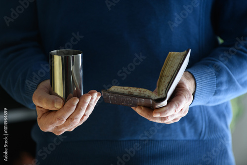 A man holds a silver cup filled with wine and recites the Jewish  blessing of Kiddush from a prayerbook.