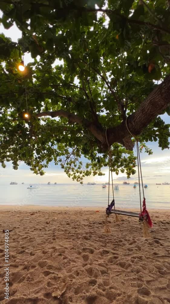 A tranquil beach scene with a swing hanging from a tree, set against the peaceful backdrop of boats on calm water during golden hour. Perfect for serene, tropical, and relaxing themes. A tranquil beach scene with a swing hanging from a tree, set against the peaceful backdrop of boats on calm water during golden hour. Perfect for serene, tropical, and relaxing themes.