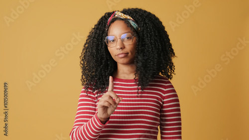 Young woman with curly hair wearing glasses wearing striped sweater, looks at the camera, listens carefully and strongly disagrees, on yellow background in the studio