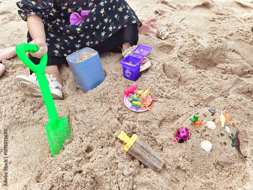 The girl is sitting on the sand and playing sand toys set. A little girl playing beach toys. Kid activity. The kid playing toys in the sandbox The girl is sitting on the sand and playing sand toys set. A little girl playing beach toys. Kid activity. The kid playing toys in the sandbox