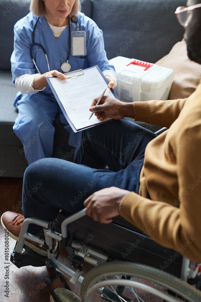 Fototapeta premium Vertical cropped shot of African American man using wheelchair and signing medical form with female doctor holding clipboard