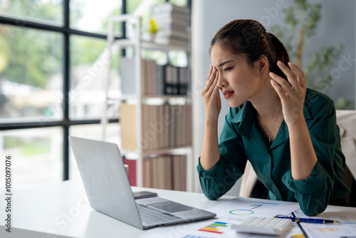 A woman is sitting at a desk with a laptop and a stack of papers