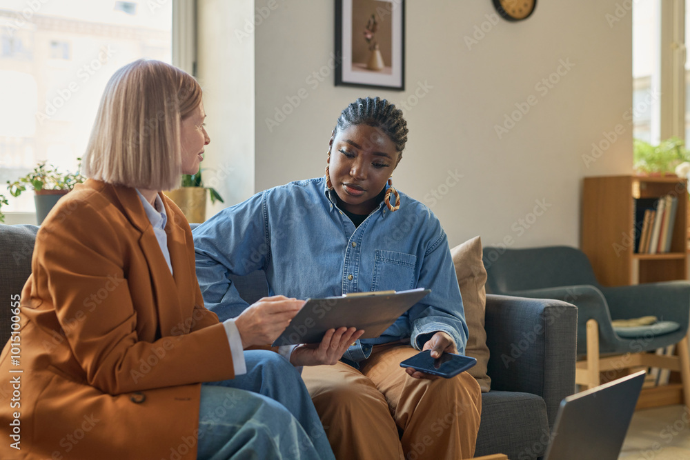 Portrait of adult African American woman talking to female finance expert explaining tax forms and showing clipboard with documents Portrait of adult African American woman talking to female finance expert explaining tax forms and showing clipboard with documents