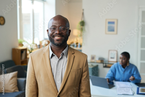Waist up portrait of adult Black man wearing glasses and looking at camera with smile standing indoors in home interior, copy space