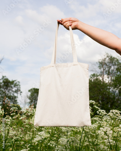 Mature woman, female hand holding white bag canvas fabric for mock up blank template on meadow with flowers background. Ecology, reusable concept. Eco friendly tote shopper