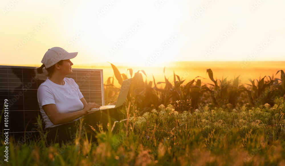 Woman farmer wearing white cap and t-shirt with laptop sits next to solar panel at sunset Woman farmer wearing white cap and t-shirt with laptop sits next to solar panel at sunset