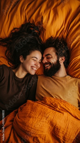 A candid moment of a couple lying on a bed covered with orange sheets, smiling at each other in a relaxed and joyful atmosphere. The warmth of the orange tones enhances the intimate connection.