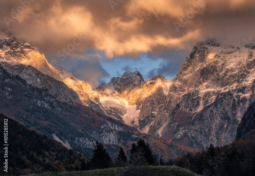 Sunlit snow-capped mountain peaks glow under dramatic clouds during sunset in autumn. Alps. Serene landscape with rocks in snow, colorful orange sky, trees in fall. Alpine mountains in Slovenia