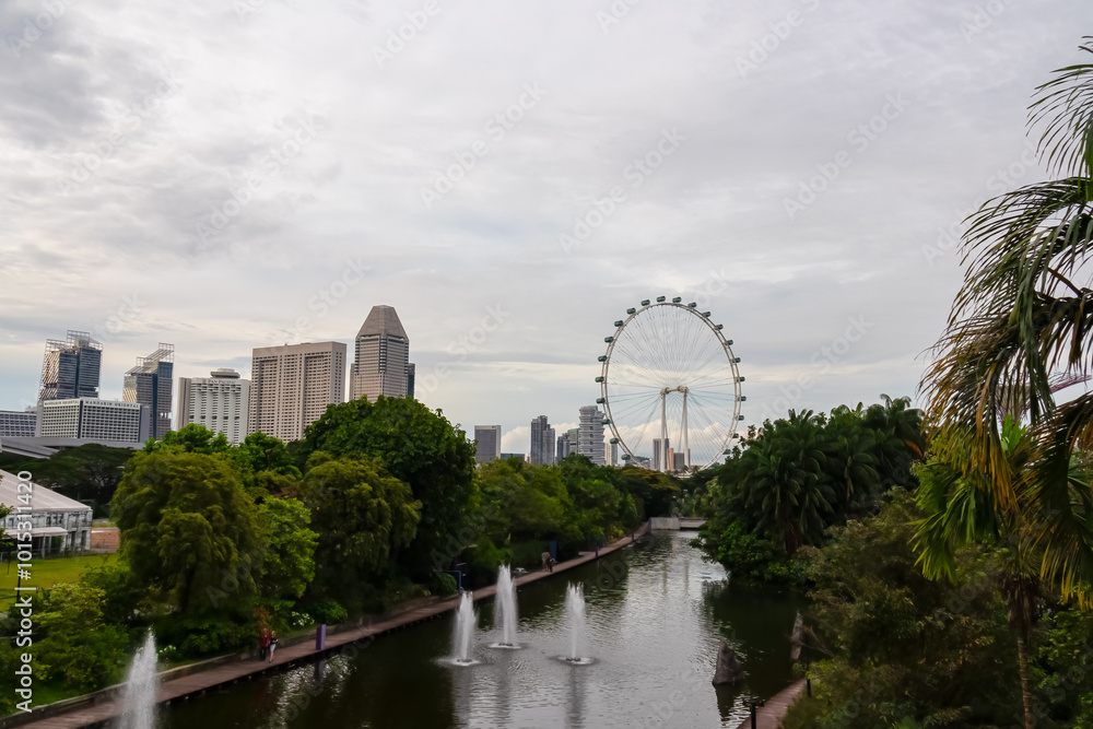 Naklejka premium Pathway winds along tranquil lake in Gardens by the Bay, Singapore. Trail is bordered by lush greenery and colorful flowers. Iconic Singapore Flyer Ferris wheel in background. Cityscape SouthEast Asia