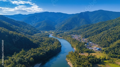 An aerial view of a winding river flowing through a valley surrounded by lush green mountains under a blue sky with white clouds.