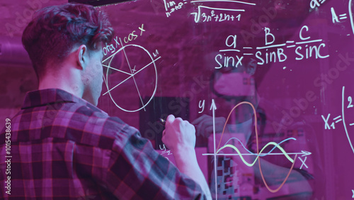 Young man solves mathematical equations on blackboard. Media. Young man solves complex equations on transparent board. Genius writes equations on blackboard in neon light