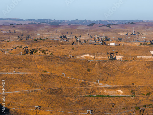 A vast desert landscape in California features numerous oil rigs amidst rolling hills. The scene includes power lines and small structures under a clear sky.