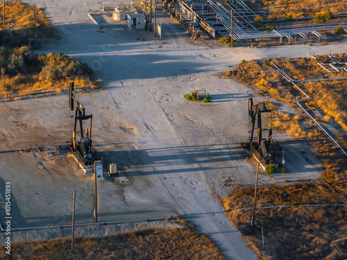 An aerial view shows two oil rigs in a California desert, surrounded by pipelines and industrial structures on dry, sandy terrain with sparse vegetation.