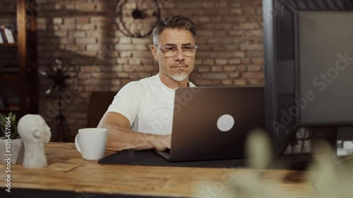 Man drinking coffee at desk in loft apartment, preparing for long business day. Businessman in casual cloth working on laptop in home office. Portrait of happy male entrepreneur smiling.