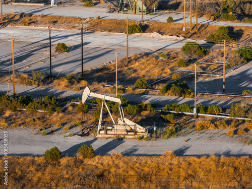 An oil rig stands in a California desert landscape, surrounded by dry grass and shrubs. Utility poles and wires indicate nearby infrastructure.