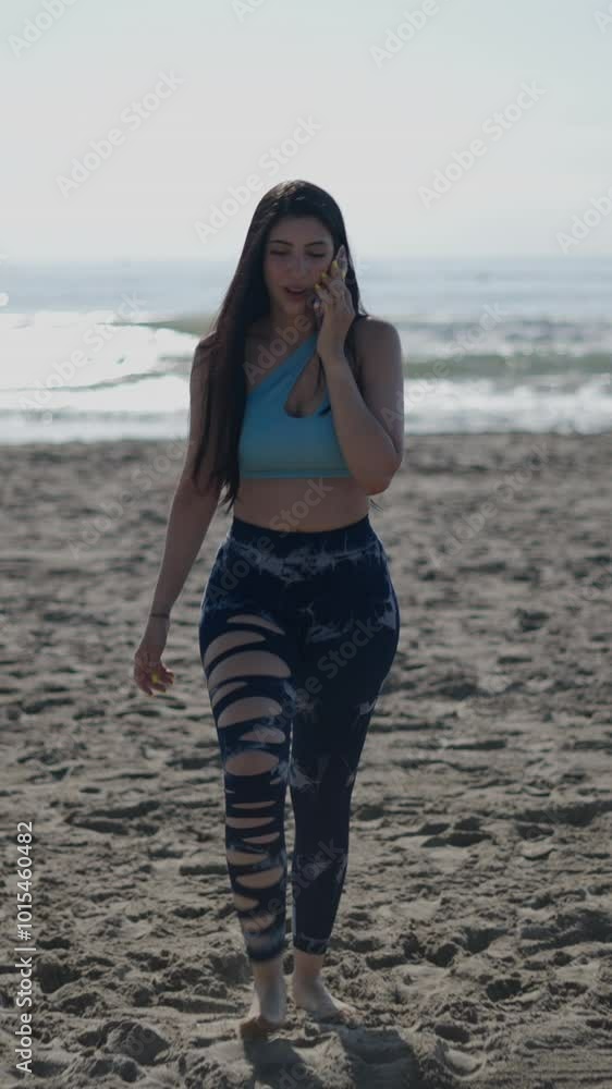 Young girl with long hair brunette Caucasian woman walking facing camera on the sand of the beach on a sunny day makes a call on her smartphone Young girl with long hair brunette Caucasian woman walking facing camera on the sand of the beach on a sunny day makes a call on her smartphone