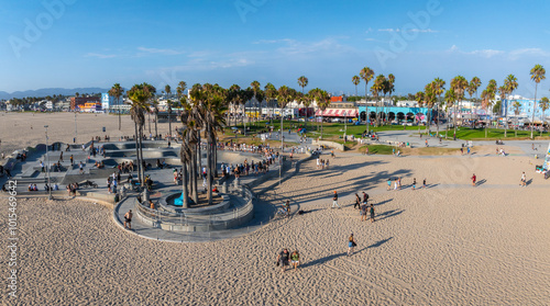 Fototapeta Naklejka Na Ścianę i Meble -  Venice Beach in Los Angeles features a skate park, sandy beach with palm trees, colorful buildings along the boardwalk, and distant mountains under a clear sky.