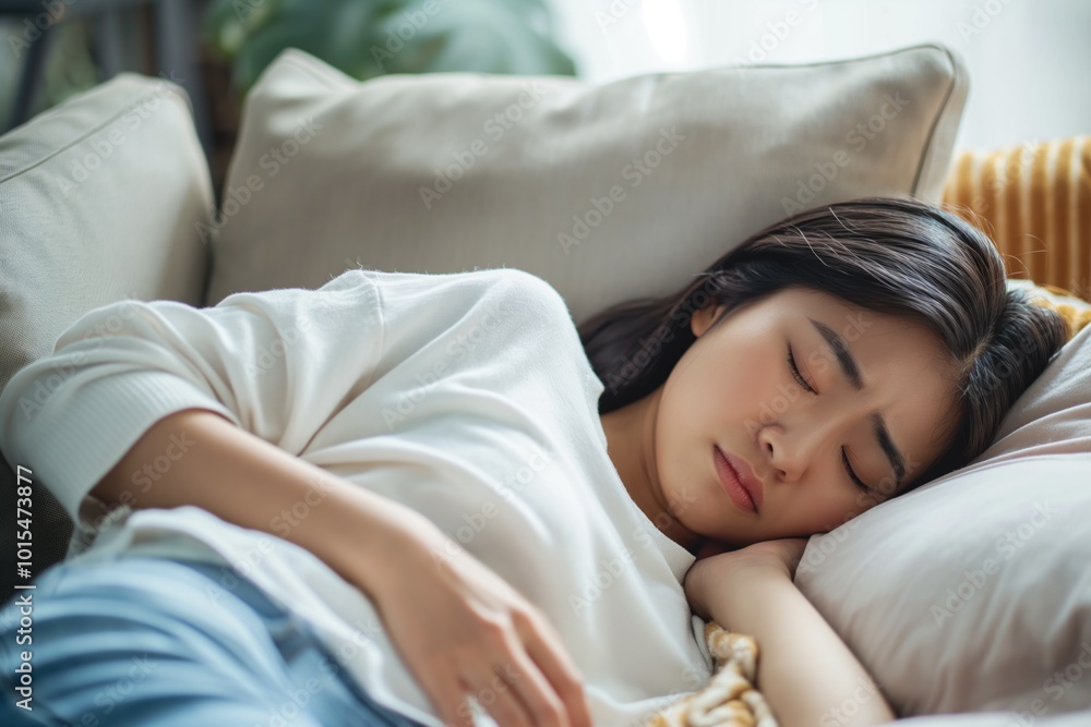 Young Asian woman lies on beige couch, resting head on white pillow. Soft fills room, creating peaceful atmosphere. Woman dressed in white shirt, blue jeans, with yellow blanket draped over pillow. Young Asian woman lies on beige couch, resting head on white pillow. Soft fills room, creating peaceful atmosphere. Woman dressed in white shirt, blue jeans, with yellow blanket draped over pillow.