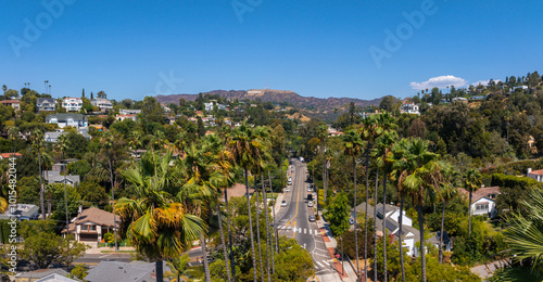 The image captures the Hollywood district in Los Angeles, featuring the Hollywood Hills and Sign. A tree lined street with palm trees and houses is visible.