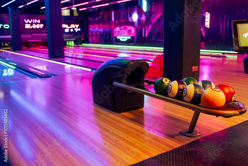 Various bowling balls arranged in stand with wooden lane at bowling hall. Illuminated play rom in shopping center with no people. 