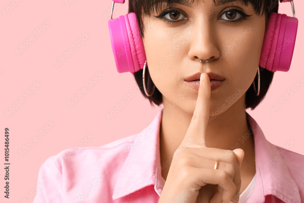 Stylish young woman in headphones with nose piercing showing silence gesture on pink background Stylish young woman in headphones with nose piercing showing silence gesture on pink background