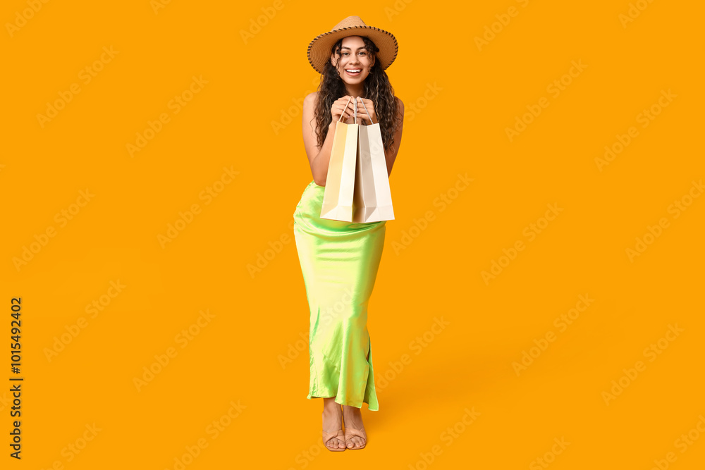 Stylish African-American woman in dress and hat holding shopping bags on yellow background Stylish African-American woman in dress and hat holding shopping bags on yellow background