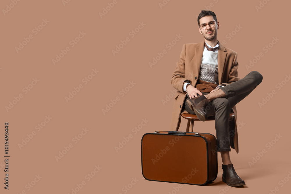 Handsome young man in stylish coat with eyeglasses and suitcase sitting on chair against beige background Handsome young man in stylish coat with eyeglasses and suitcase sitting on chair against beige background