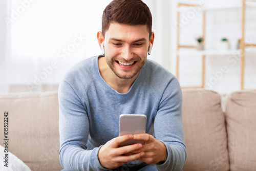 Mobile User. Joyful Man In Wirelss Earphones Using Smatphone Playing Games Sitting On Sofa Indoor. photo