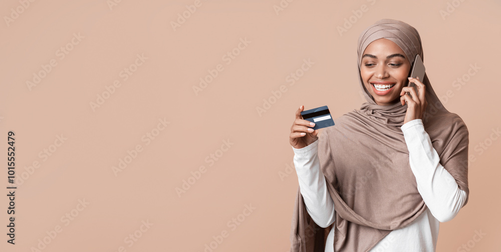Fototapeta premium Customer service concept. Young black muslim woman in hijab holding credit card and talking on cellphone with bank manager, yellow background