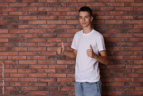 Teenage boy wearing white t-shirt and showing thumbs up near brick wall, space for text