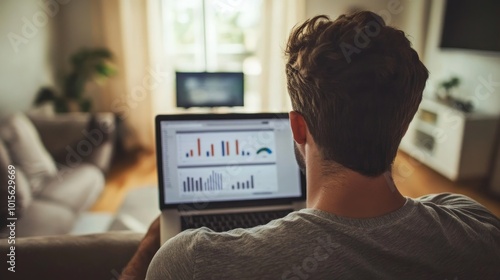 A man sits on a couch in a living room, looking at a laptop screen with graphs and charts.