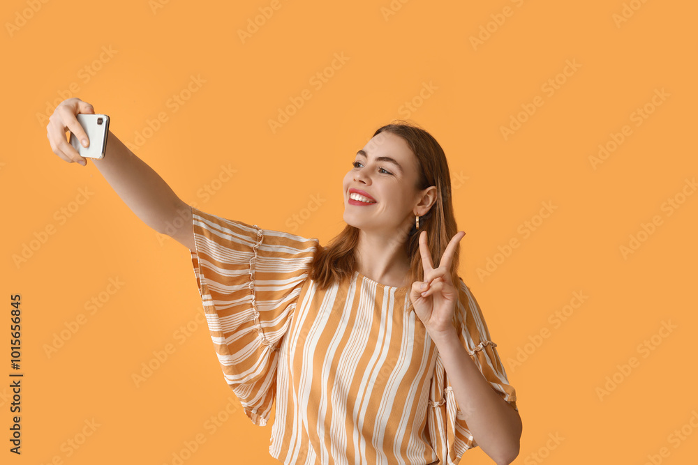Fototapeta premium Pretty young woman showing peace gesture while taking selfie on orange background