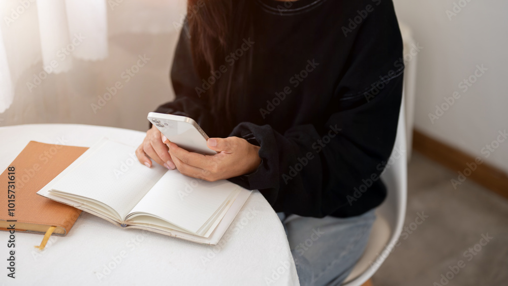 Obraz premium A close-up of a woman in a cozy sweater using her smartphone while reading at a table indoors.