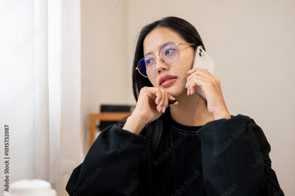A beautiful, thoughtful Asian woman is having a conversation on the phone while sitting indoors. A beautiful, thoughtful Asian woman is having a conversation on the phone while sitting indoors.