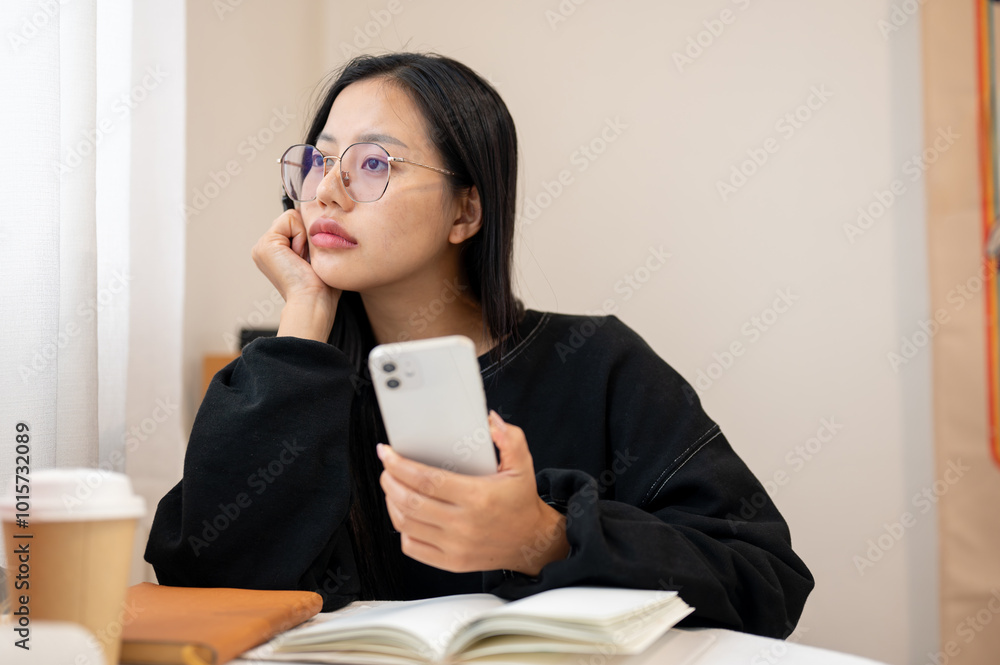 A thoughtful Asian woman gazes out the window while sitting at an indoor table, deep in thought. A thoughtful Asian woman gazes out the window while sitting at an indoor table, deep in thought.