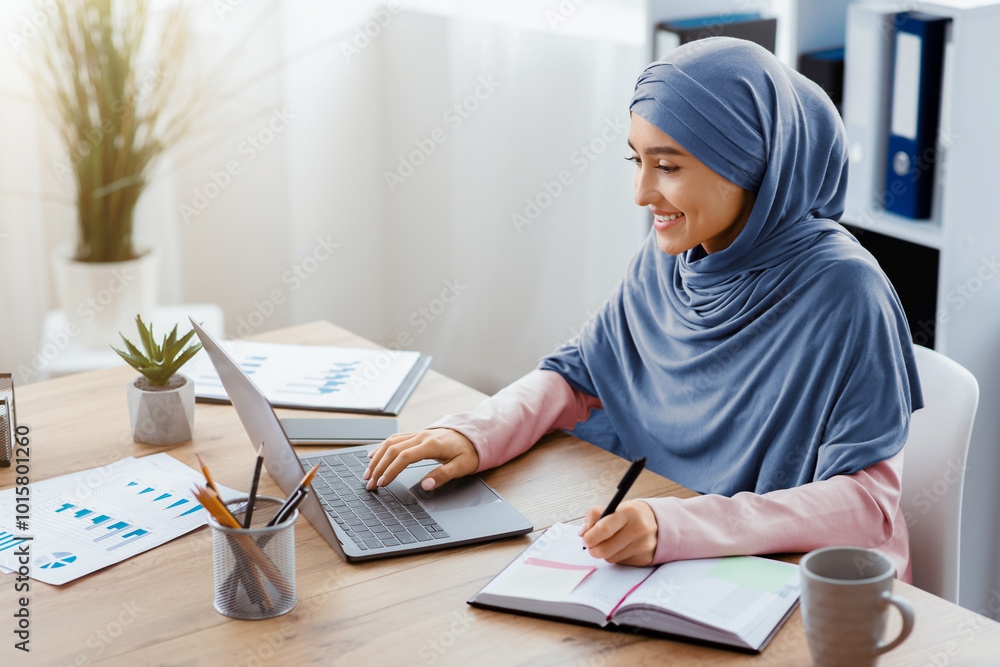 Muslim woman in hijab using laptop and taking notes in modern office, islamic female employee sitting at workplace, side view with copy space Muslim woman in hijab using laptop and taking notes in modern office, islamic female employee sitting at workplace, side view with copy space