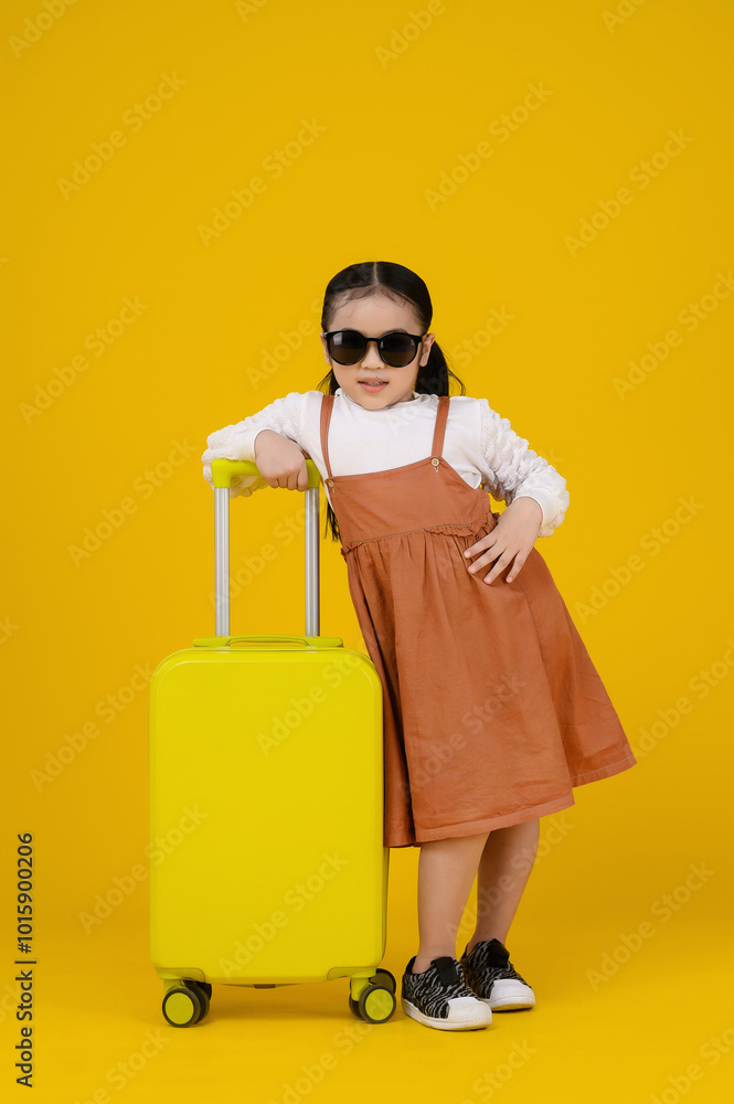 Asian confident girl kids wearing sunglasses and a brown dress leans against a yellow suitcase, posing stylishly against a vibrant yellow background. Asian confident girl kids wearing sunglasses and a brown dress leans against a yellow suitcase, posing stylishly against a vibrant yellow background.