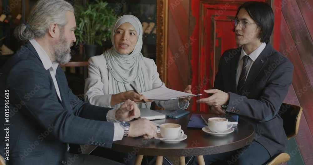 Businessmen Caucasian and Asian signing deal and talking while Middle Eastern lady in hijab interpreter translating talks in cafe. Agreement and contract concept. Businessmen Caucasian and Asian signing deal and talking while Middle Eastern lady in hijab interpreter translating talks in cafe. Agreement and contract concept.