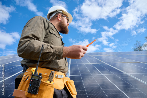 Engineer in uniform with protective helmet near solar station holding screwdriver. Supervisor is checking on solar panels on solar farm. Green energy concept. Close up low angle view.