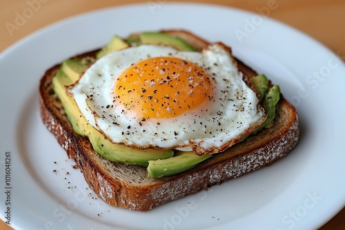 A delicious breakfast toast topped with avocado and a sunny-side-up egg, seasoned with black pepper on a white plate.