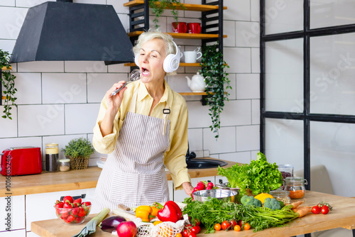 happy beautiful elderly gray haired senior woman in headphones cook in cozy kitchen with fresh organic vegetables, having fun and singing