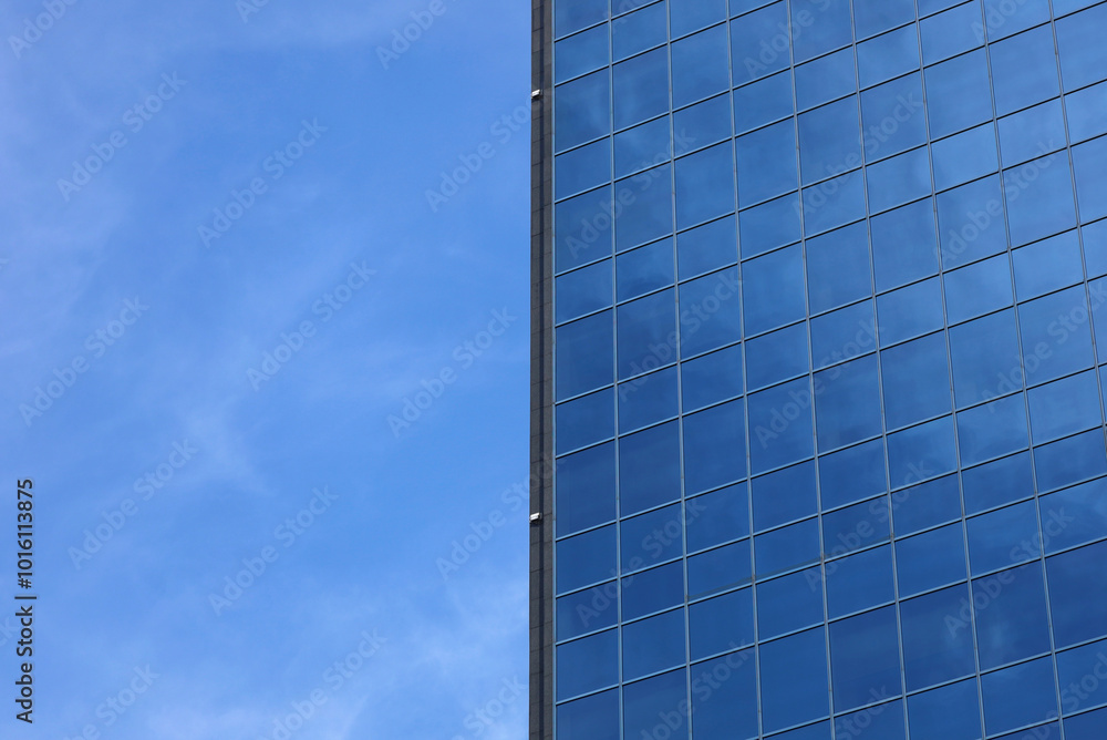 Image is divided into two halves. The left half is blue sky and right half is modern office building facade made of blue toned windows. Fragment of skyscraper. Place for text.