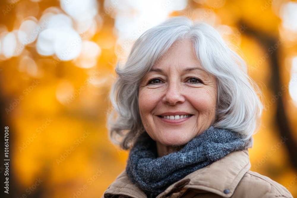 An elderly woman with a gentle smile and gray hair is pictured wearing a coat and scarf in a beautifully lit autumn scene, surrounded by glowing orange foliage. An elderly woman with a gentle smile and gray hair is pictured wearing a coat and scarf in a beautifully lit autumn scene, surrounded by glowing orange foliage.
