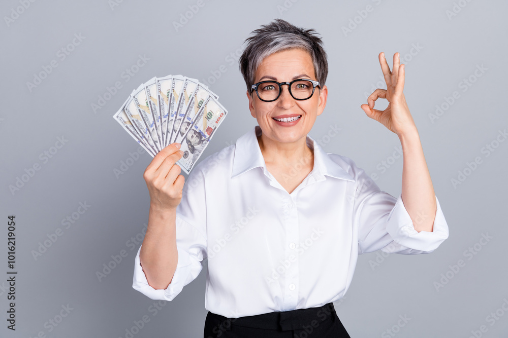 Photo portrait of lovely senior lady hold money fan okey approve dressed stylish formalwear isolated on gray color background Photo portrait of lovely senior lady hold money fan okey approve dressed stylish formalwear isolated on gray color background