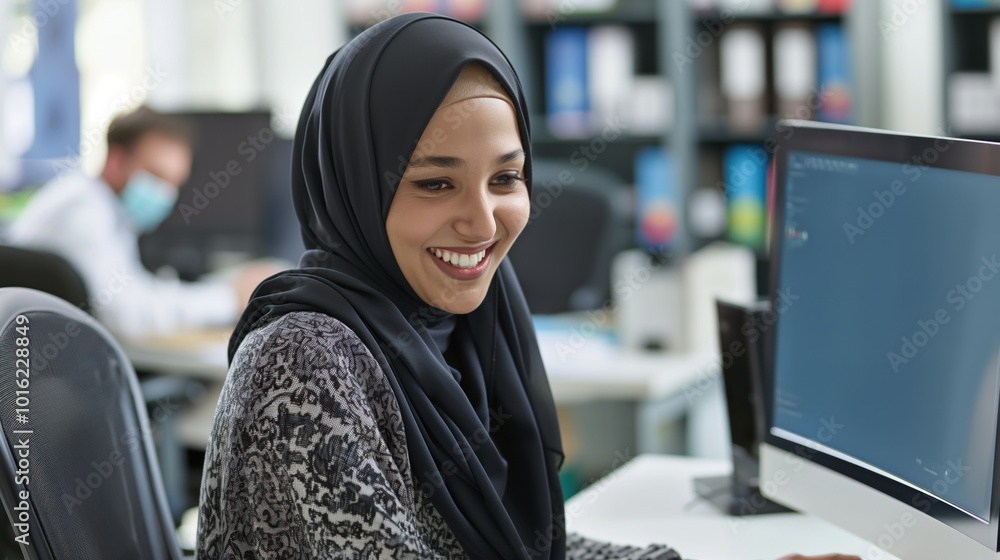 Woman works on computer in modern office. She wears black hijab, gray sweater, focuses on laptop screen. Coffee cup, bookshelf in background. Desk, keyboard, monitor. Woman works on computer in modern office. She wears black hijab, gray sweater, focuses on laptop screen. Coffee cup, bookshelf in background. Desk, keyboard, monitor.