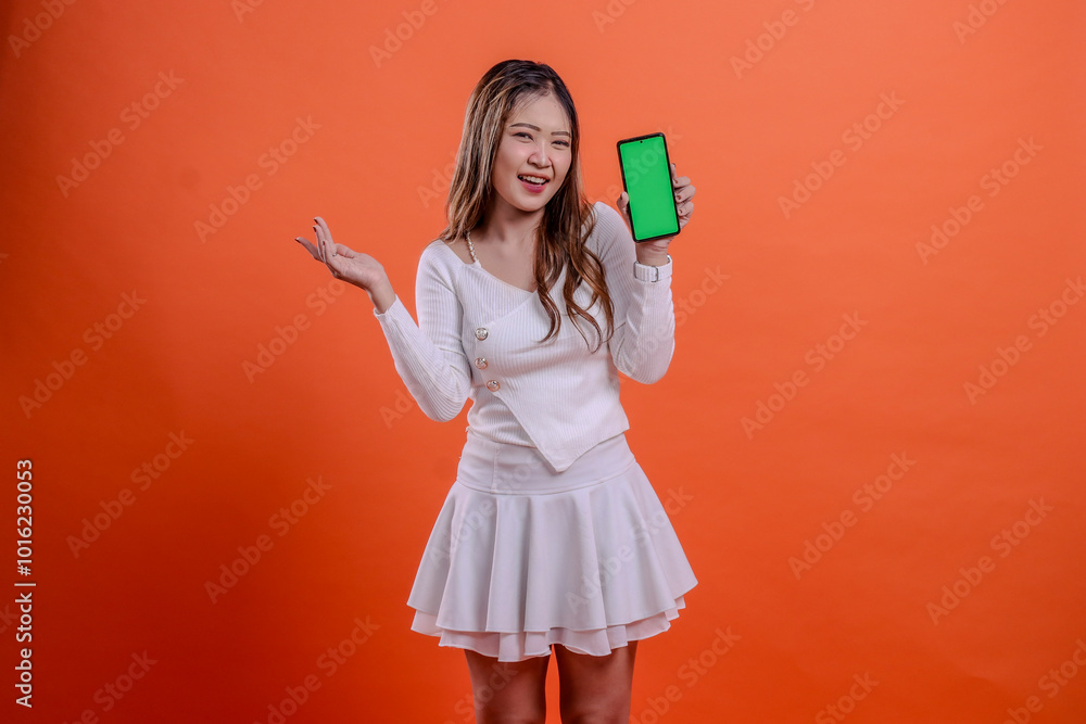 expression of beautiful adult asian woman in white top standing cheerfully to camera hand holding mobile phone screen, cellphone while presenting to right isolated orange background. for technology expression of beautiful adult asian woman in white top standing cheerfully to camera hand holding mobile phone screen, cellphone while presenting to right isolated orange background. for technology