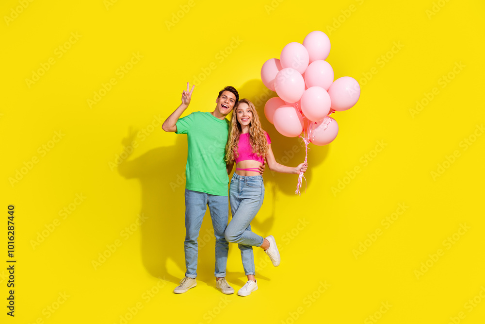Full size photo of two nice young people show v-sign hold balloons isolated on bright yellow color background Full size photo of two nice young people show v-sign hold balloons isolated on bright yellow color background