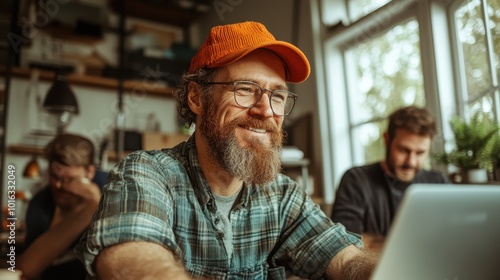 A cheerful worker wearing an orange cap enjoys working on a technology project with a laptop, surrounded by colleagues, conveying collaboration and innovation.